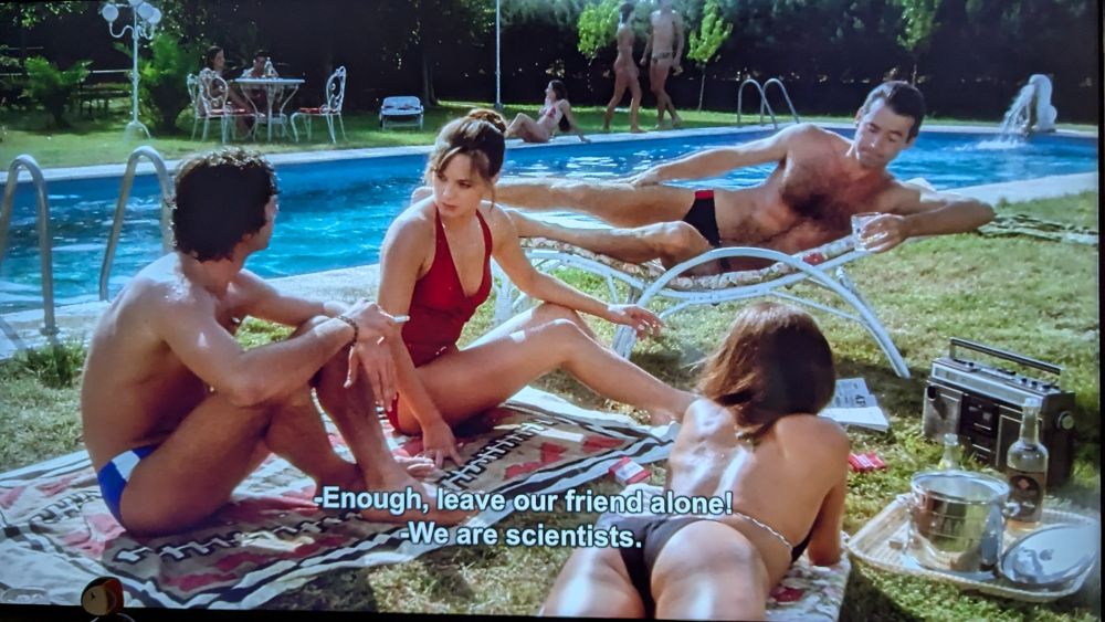 Two men and a woman are sitting beside a pool. One woman is saying "enough, leave our friend alone! We are scientists."