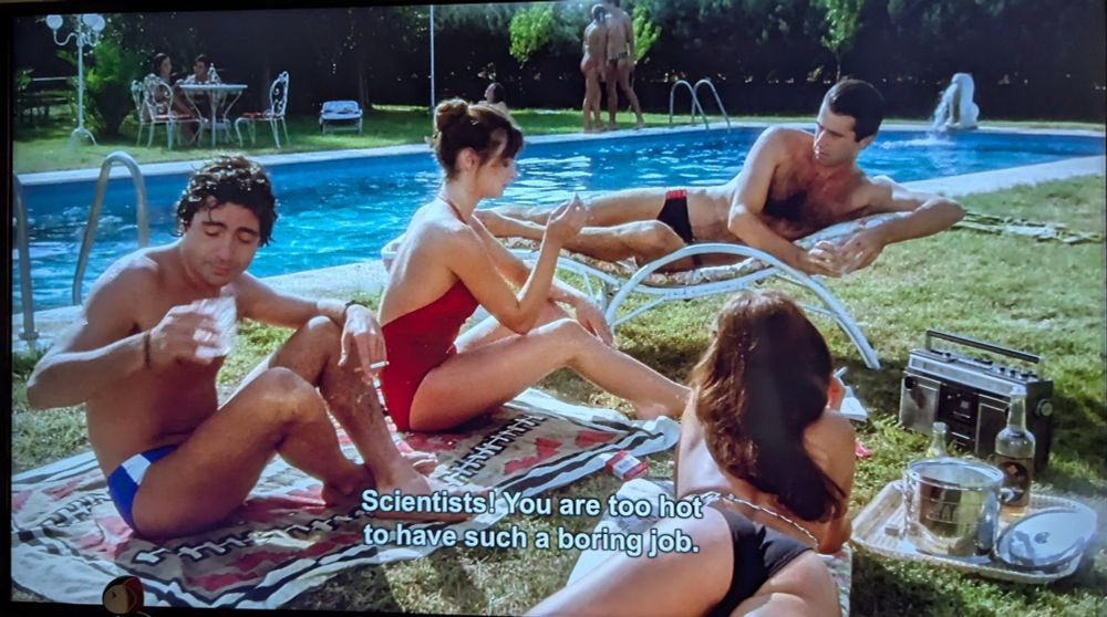 Two men and a woman are sitting beside a pool. One man is saying "scientists! You are too hot to have such a boring job."