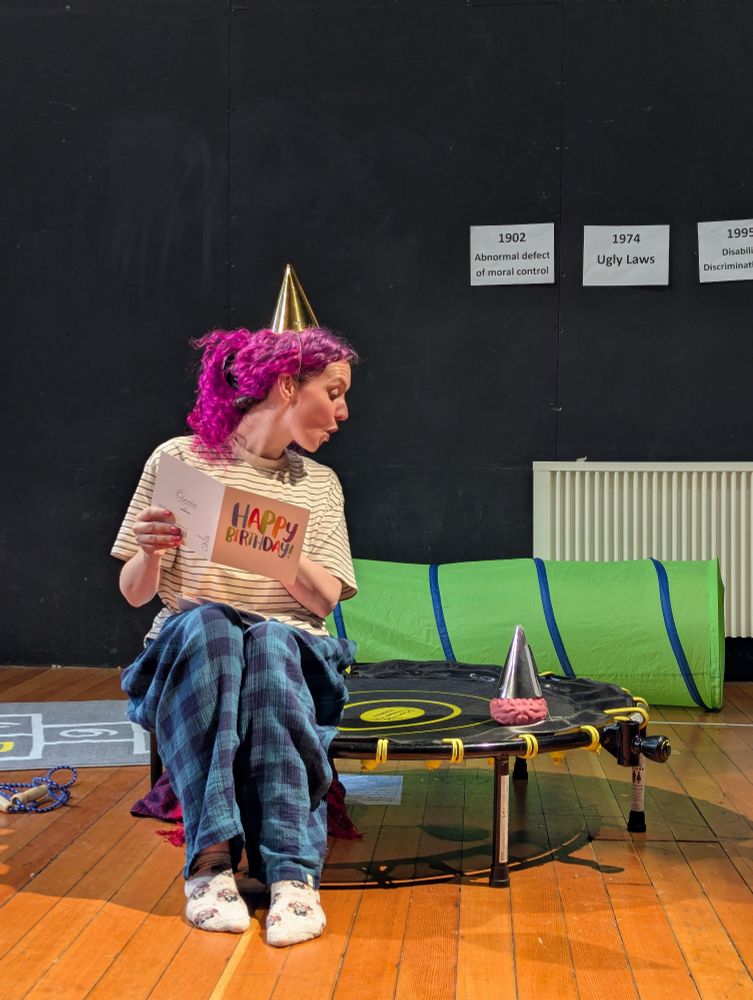 A rehearsal shot from Fiona Moon's show, A Brief History of Neurodivergence. Fiona, a purple-haired white woman wearing a party hat, sits on a trampoline reading a birthday card to a pink squishy brain which is also wearing a party hat. 