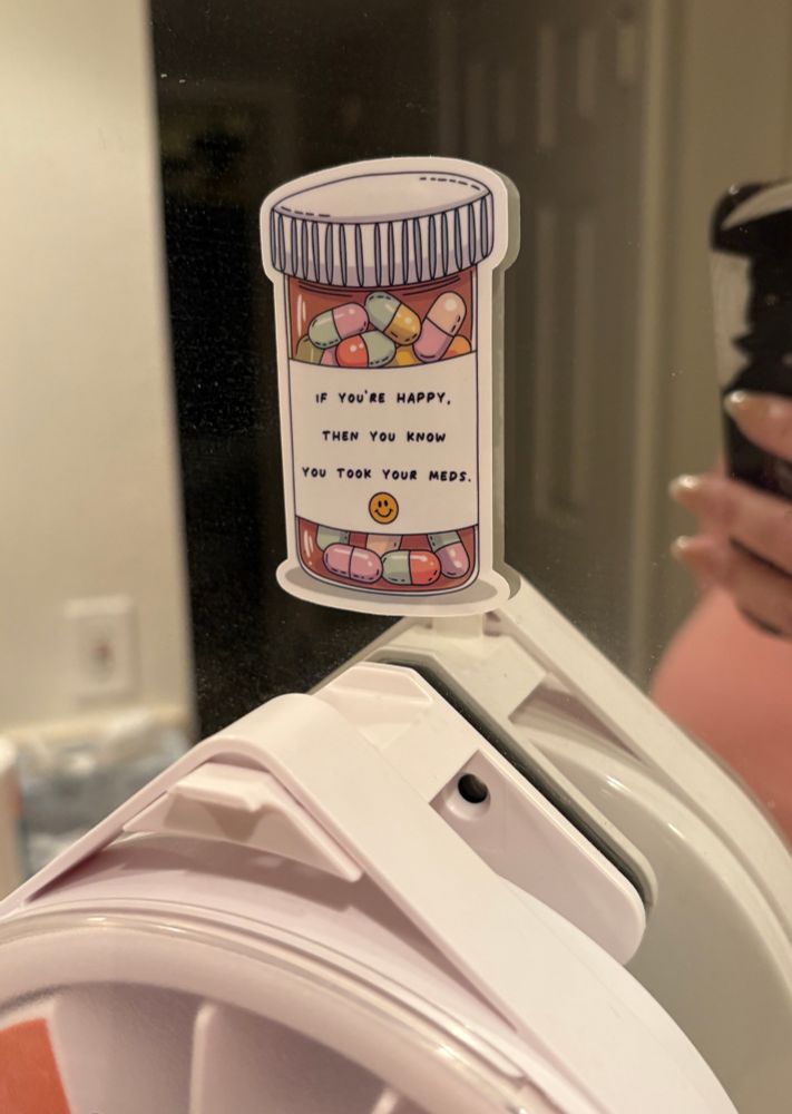 Sticker of a pill bottle full of multi-colored capsules with a label that reads “If you’re happy then you know you took your meds”. Sticker is on a mirror above a pill dispenser. 