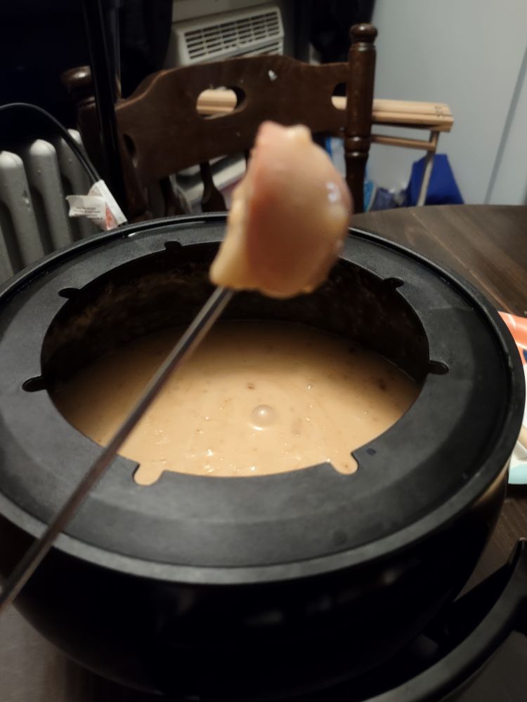 An electric fondue pot with melted cheese and a skewer with a piece of pretzel bun coated in cheese.