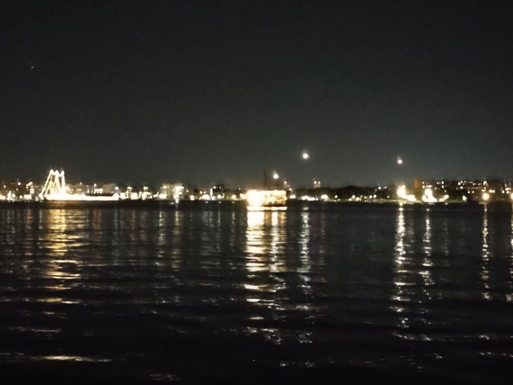 Boston Harbor at night. The sky is dark, the golden light of the city are reflected in black, rippling water. The glowing lights of Logan Airport are in the background with some white dots in the air of planes lining up for landing.