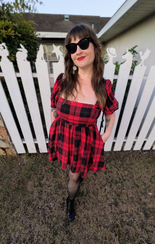 A woman is posing in front of a white fence modeling a dress she sewed herself. The dress has a low cut, square neck, empire waist bodice with puffed sleeves, and is paired with a simple gathered skirt with pockets. The dress is made with red and black buffalo check plaid in a cotton blend, and it's styled with gold earrings, a gold necklace, black sunglasses, black boots, and black tights. 