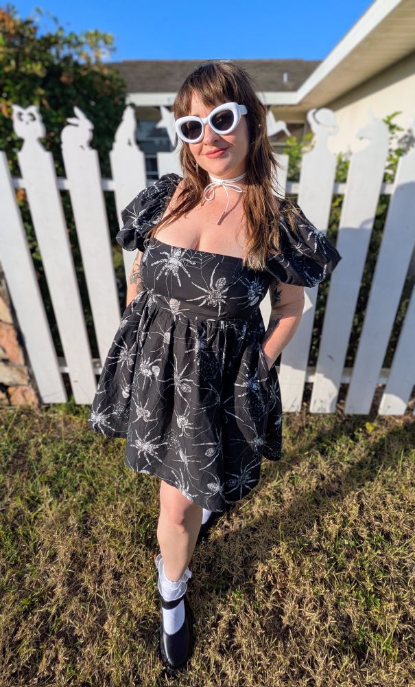 A woman is posing in front of a white fence wearing a dress she sewed herself. The dress has a square neck, low cut, empire waist bodice with short puffed sleeves, and is paired with a gathered skirt. The dress is made with a black cotton voile fabric patterned with large grey and white spiders. The look is styled with white sunglasses, white spiderweb earrings, a white choker necklace, white lacy crew cut socks, and black Mary Jane shoes. 