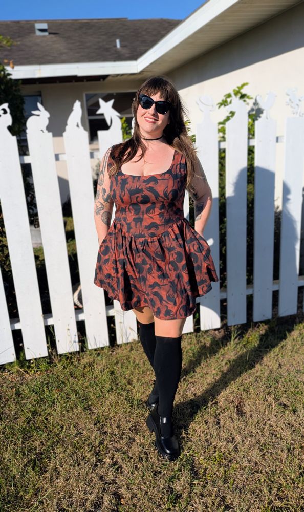 A woman is posing in front of a white fence wearing a dress she sewed herself. The dress has a sleeveless princess seam bodice with a scooped neckline paired with a gathered at the waist short skirt with pockets. The dress is made with red cotton poplin patterned with black cats. The outfit is styled with a plain black choker necklace, black cat earrings, knee high black stockings, and Mary Jane shoes. 