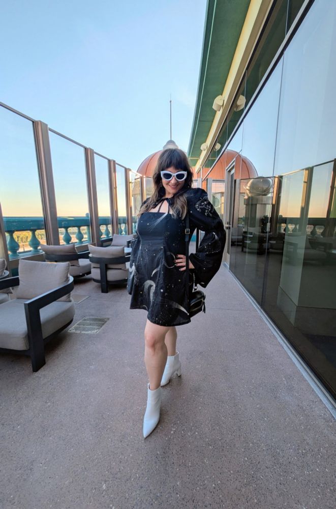 A woman is posing at a rooftop bar modeling a dress she sewed herself. The dress features a tie front keyhole neck with gathered long bell sleeves and an A-line short skirt. The dress is made with black cotton voile patterned with white and gold celestial objects, such as comets, constellations, and stars. The dress is styled with white ankle boots and white sunglasses. 