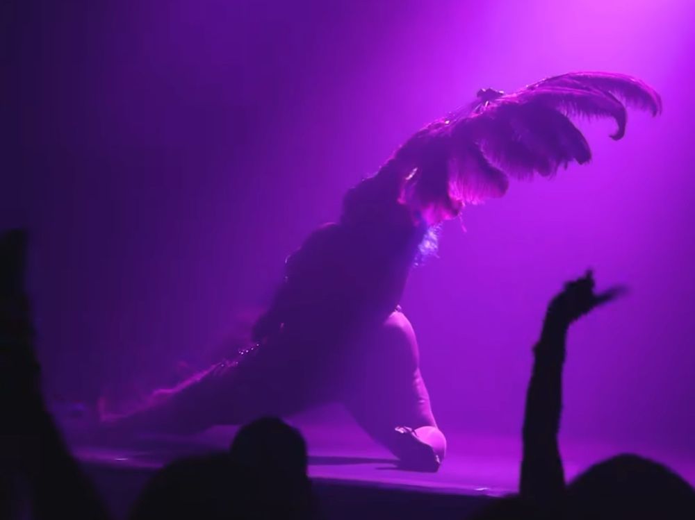 Bianca, as Roxy, bathed in pink light posed on the ground with feather fans