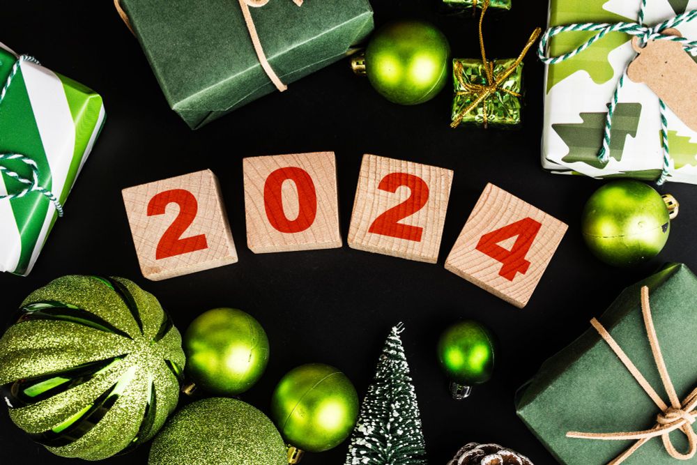 Wooden blocks writing out 2024 are scattered across a black table cloth. They are surrounded by green and shiny baubles and gifts, giving a festive air. 