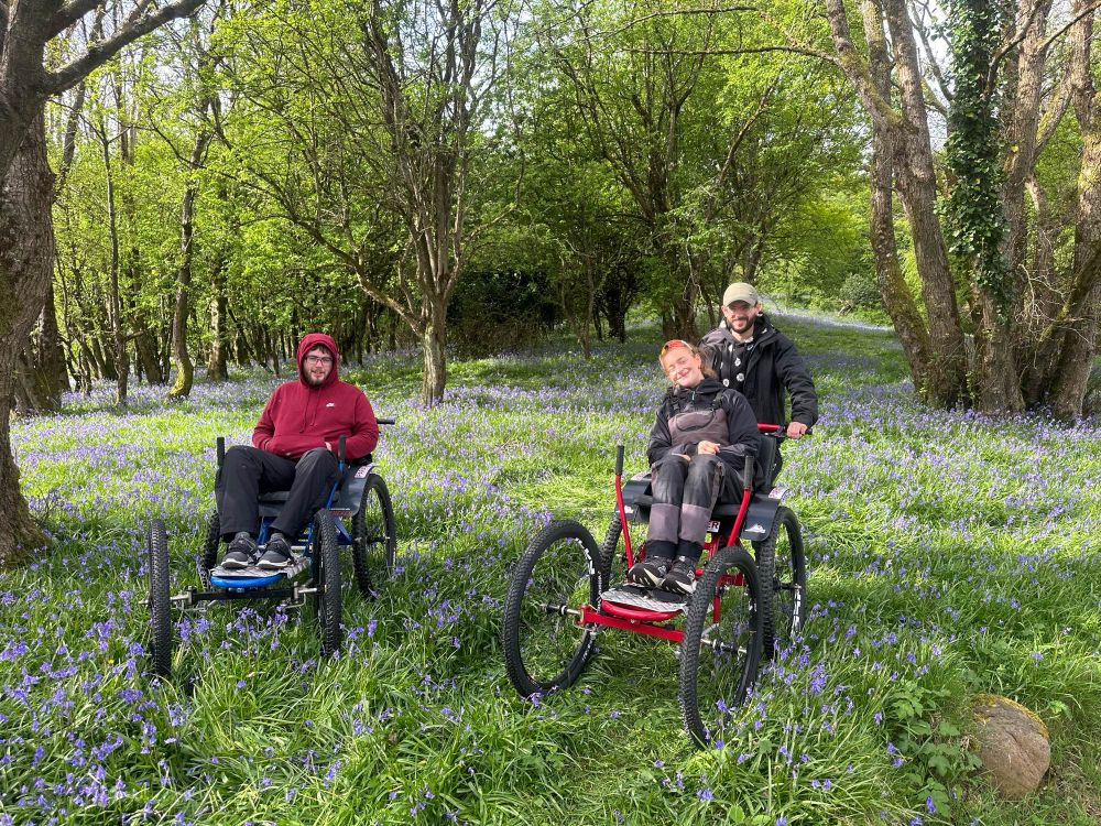 Two wheelchair users in a pair of specialist 4 wheeler off road wheelchairs stopped amidst woodland and a carpet of bluebells 