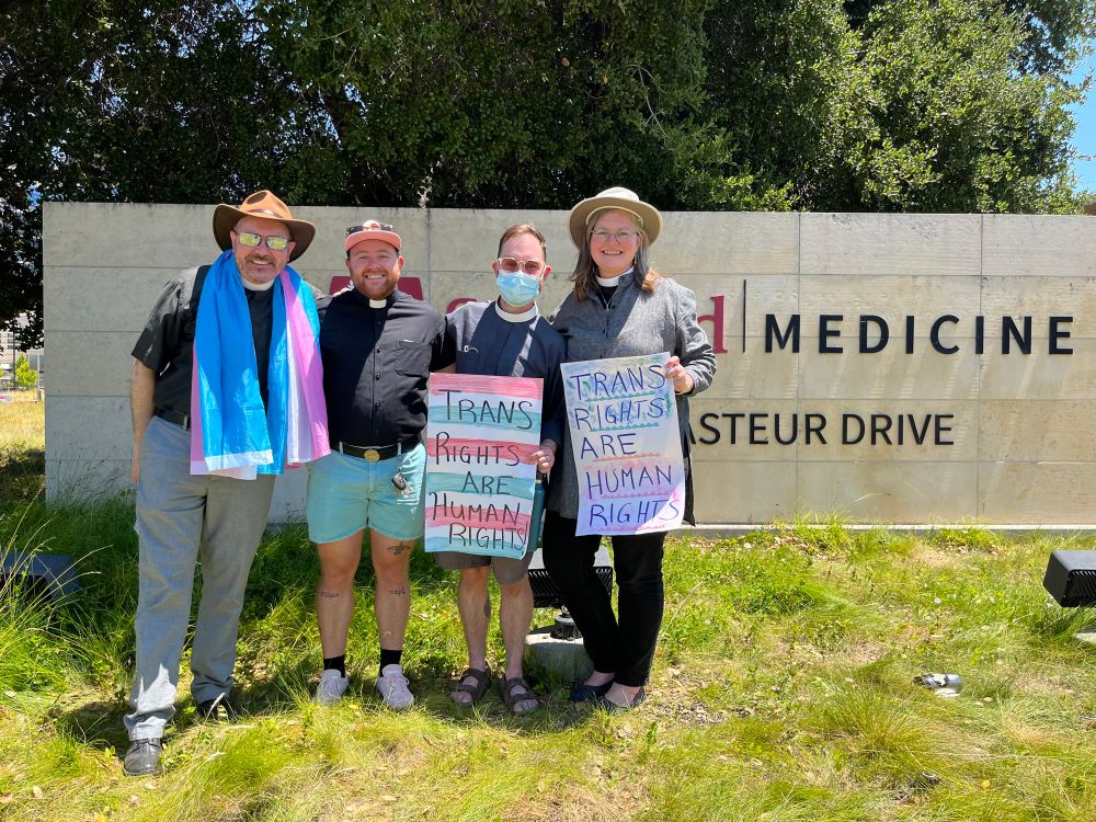 Trans and allied Episcopal clergy rally outside Stanford Medical Center. There are four adults in the photo, all wearing clerical collars; two are holding hand-lettered signs in pink and blue that say “Trans Rights Are Human Rights.” 