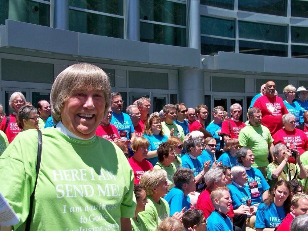 Deacon Vicki Gray stands, smiling broadly, amidst a crowd of fellow advocates for LGBTQ inclusion at the General Convention of the Episcopal Church in Anaheim in 2009. Vicki is wearing a bright green shirt with white lettering that says: 

HERE I AM 
SEND ME!
I am a witness 
to God’s
inclusive love 

The others in crowd are wearing similar tshirts in bright green, red, and blue colors. 