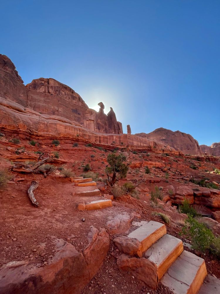 A stairway made of stone going up bright red rocks, some of the rock peaks are seen against a blue sky with the sun peaking over them
