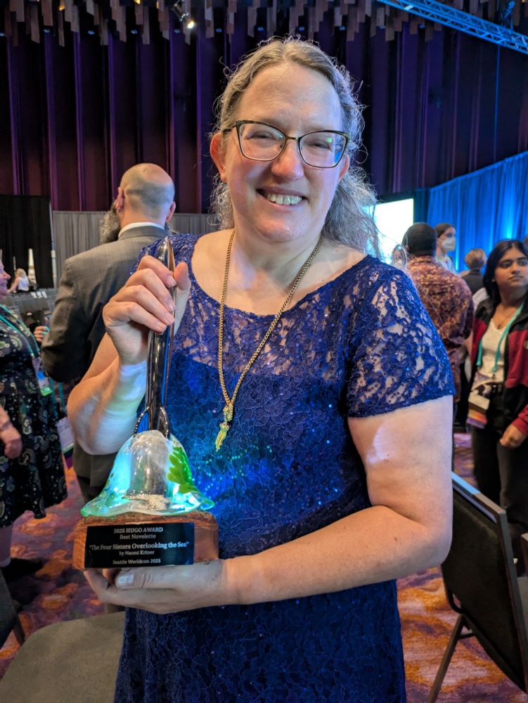 Me, in a blue dress, holding a Hugo Award trophy with a base that includes a blown glass embellishment that lights up and glows green.