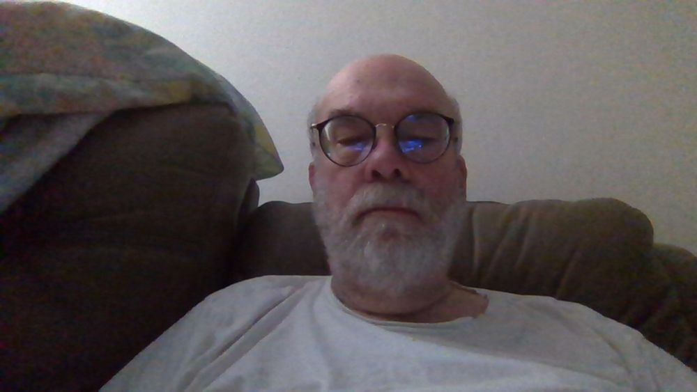 A picture of a mid-60s bald white male with glasses and a grey mustache and beard wearing a white tee shirt seated in a reclining couch.