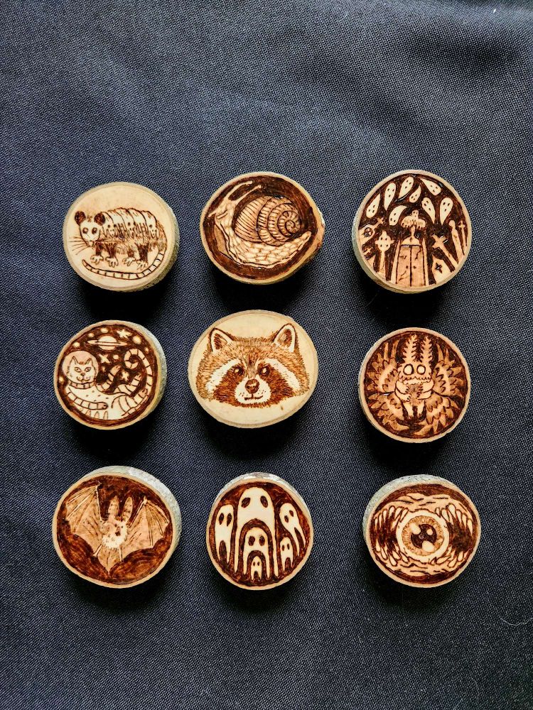 9 small wooden tokens.  From left to right top to bottom: possum with babies on back, a snail, a graveyard with ghosts, a cat in a spacesuit, a raccoon head, mothman, a bat, Fresno nightcrawlers, a monster with spikes around an eye.