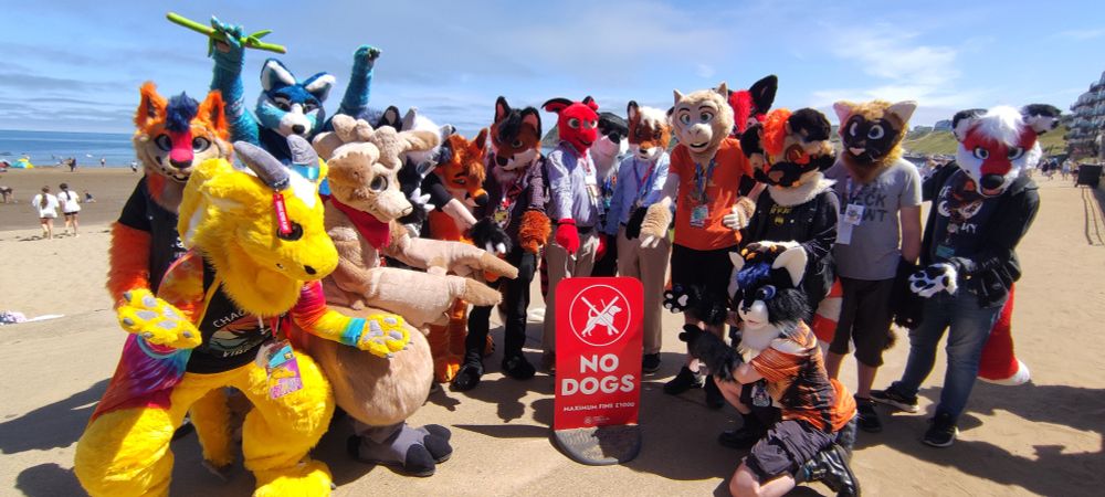 A selection of (non-canine) fursuiters point and laugh at a "no dogs" sign on the beach 