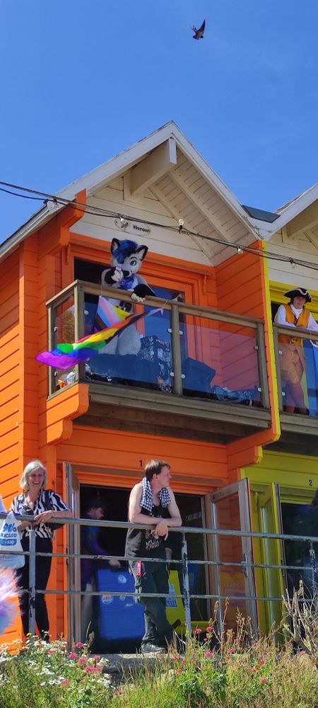 Riyo stands on a balcony above an LGBTQ flag, pointing at the viewer 