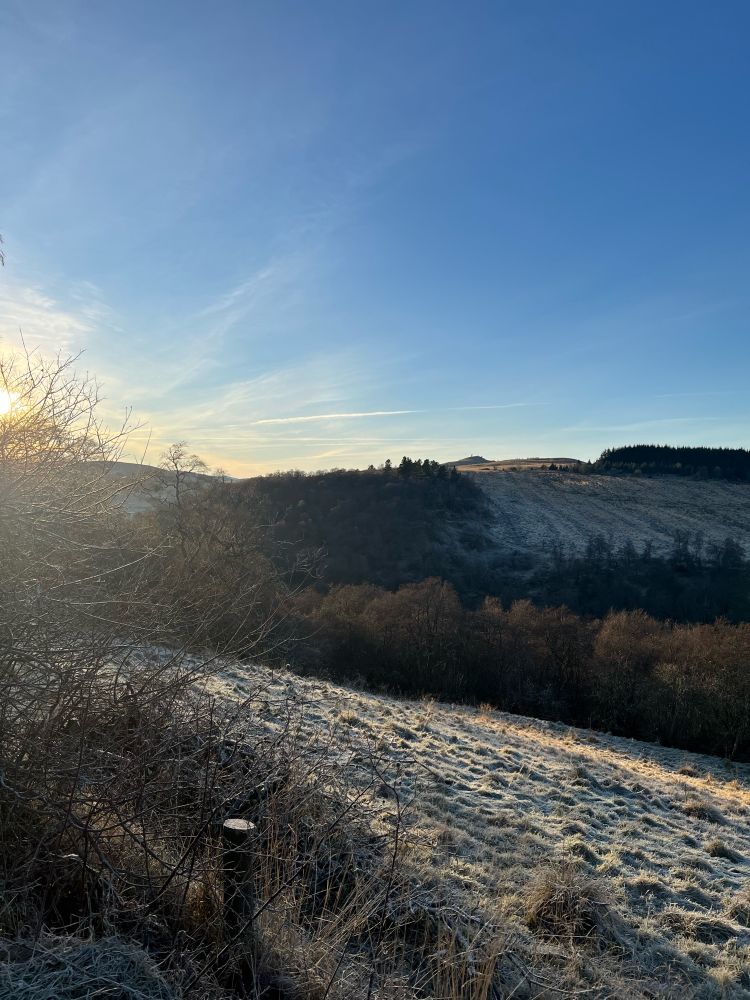 Frosty, sloping field in the foreground with a view across wooded valley and rolling hills. 