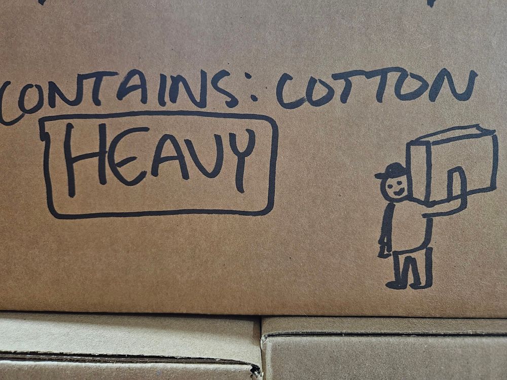simple marker drawing on the side of a box. a figure wearing a hat with a big box on their shoulder. box reads: contains: cotton HEAVY