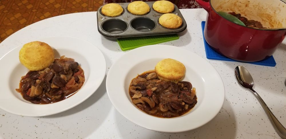 Two bowls on a kitchen counter, holding a stew made with mottled brown lima beans, mushrooms, and a tomato-based sauce, sitting next to a small red dutch oven and a pan of cornbread muffins.
