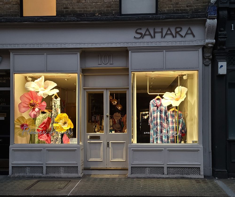 Huge paper flowers and print dresses in a shop window at night.