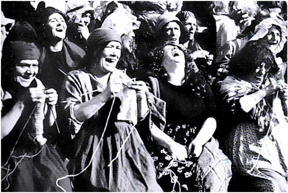 Still from a black & white movie -- poor women knitting and laughing as they watch French aristocrats being beheaded during the French Revolution. 
