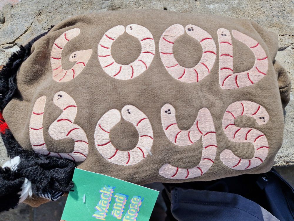 An olive green sweatshirt, folded up. There is a large embroidered design on it of the words "GOOD BOYS" spelled out by light pink cartoon worms. A small cardboard tag has been attached that says "Mack and Rose".