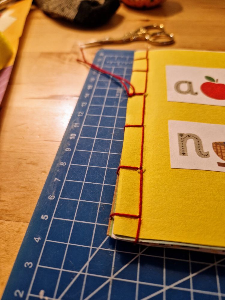 A close up of a bright yellow paper notebook with a sewn red binding in a Japanese style. The front is decorated with some illustrated alphabet letters.