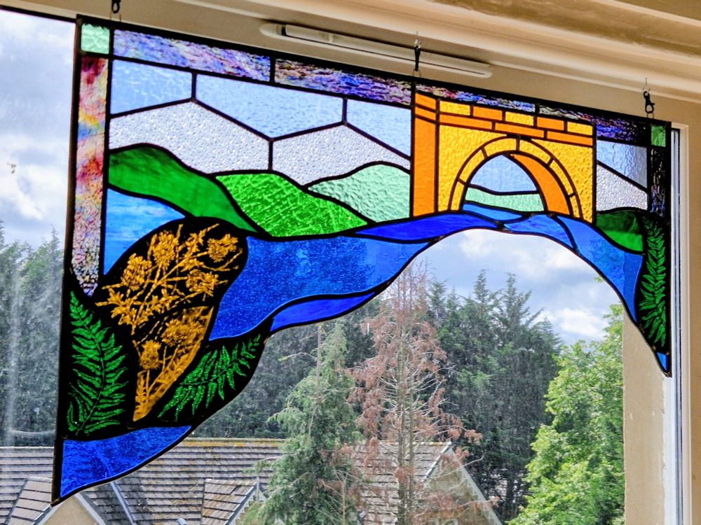 An asymmetric stained glass panel of the Water of Leith. Painted panes show ferns and thistle heads. The water flows along under a stylized bridge with hills in the background.