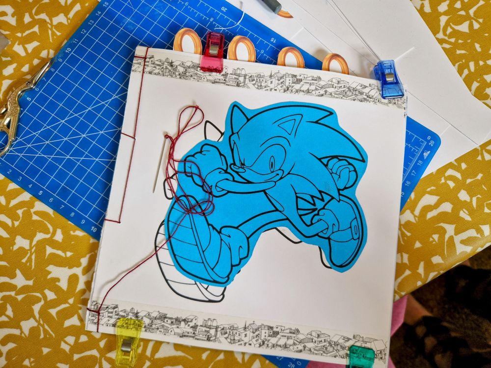 A handmade notebook in the process of being bound with dark red thread. The cover has a stylized image of Sonic the Hedgehog, blocked in by two bands of washi tape with a drawing of city rooftops. Some dividers in the shape of Sonic's rings are sticking out of the top.