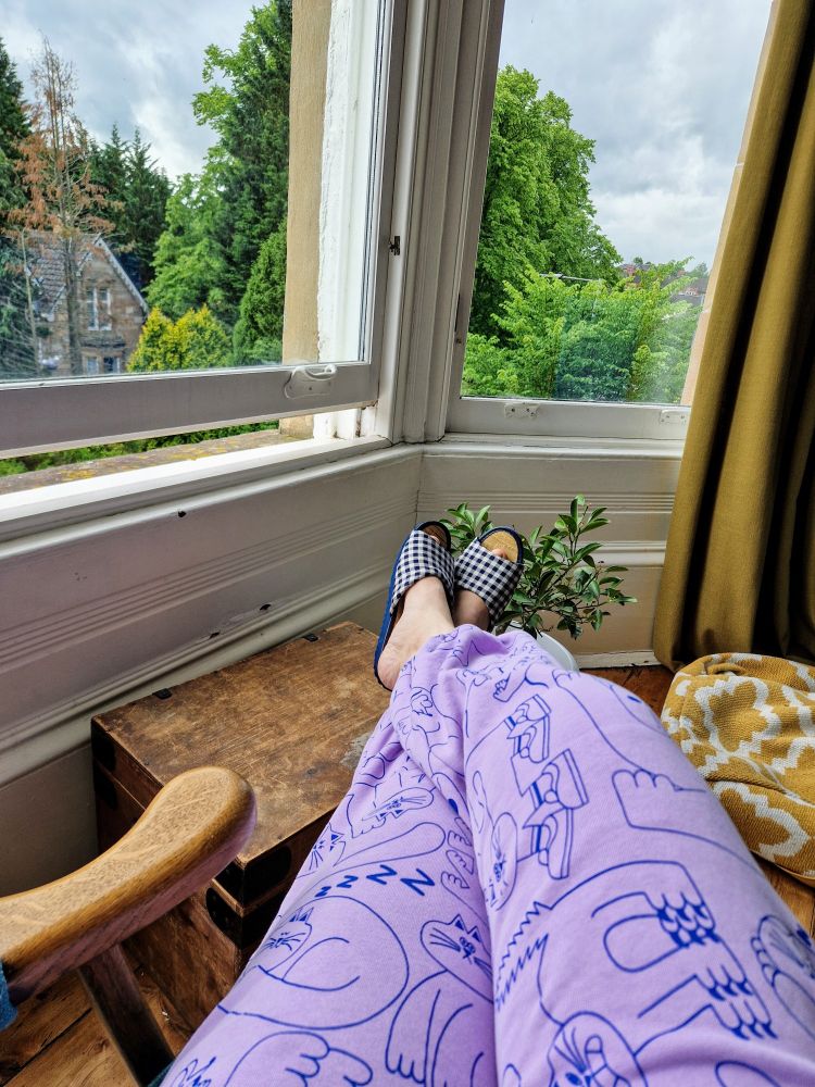 A view of a woman's legs clad in a brightly printed lilac and blue tracksuit with a cartoon cat pattern. Her feet are crossed and resting on top of an old wooden box. In the background is a peaceful view of the top of an old stone house and trees out the windows.