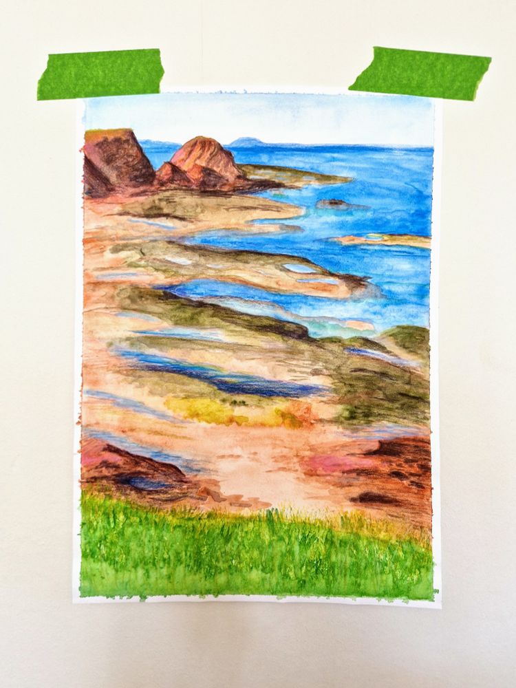 A watercolour painting of the East Lothian coastline in Scotland. Sections have been highlighted in coloured pencil. The scene shows brownish red crags fading to a flat beach, with seaweed and rock pools. A bright green grassy hill is in the foreground.