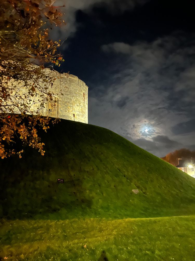 Arty nighttime photo of Clifford’s tower, a medieval limestone tower on a grassy mound against a night sky with a bright half moon 