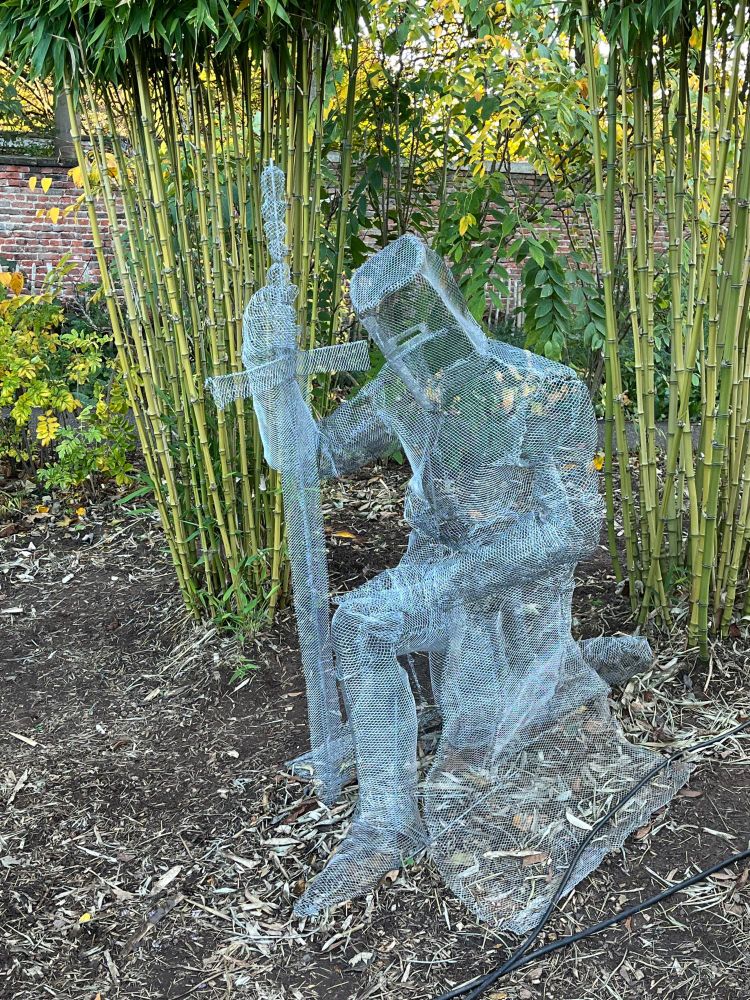 A chicken wire figure of crusader knight kneeling before his sword, as if taking an oath. Oddly, he is in several clumps of bamboo. 