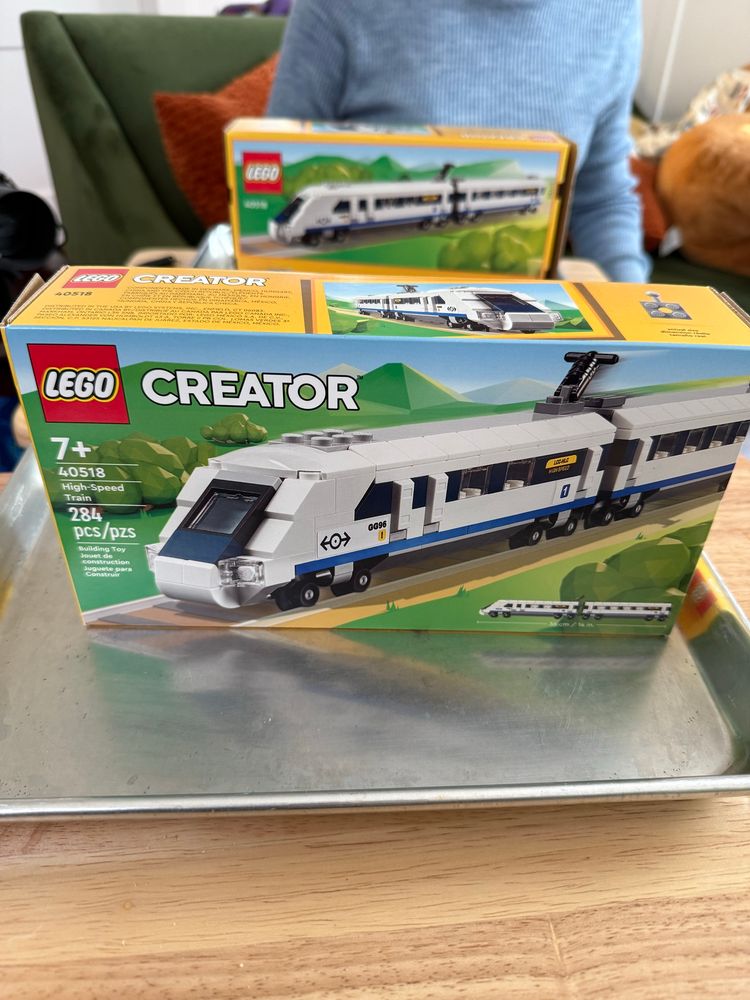 Two tray tables set up apart from each other, each has an unopened lego creator high speed train set on it. it’s a race  