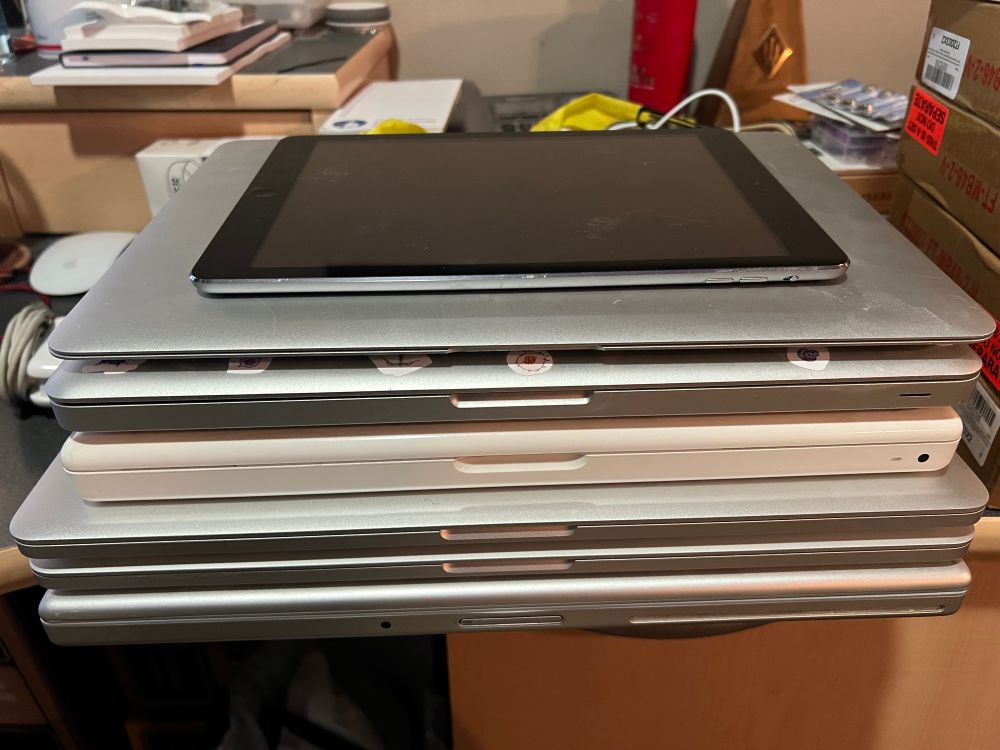 A stack of 6 MacBooks and MacBook Pros, plus an iPad on top, all sitting on my cluttered desk.