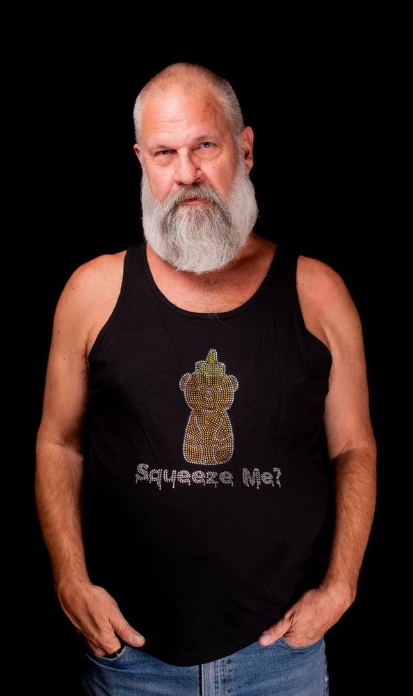 Handsome bearded daddy wearing a rhinestone tank top that shows a honey bear and the words “squeeze me?”