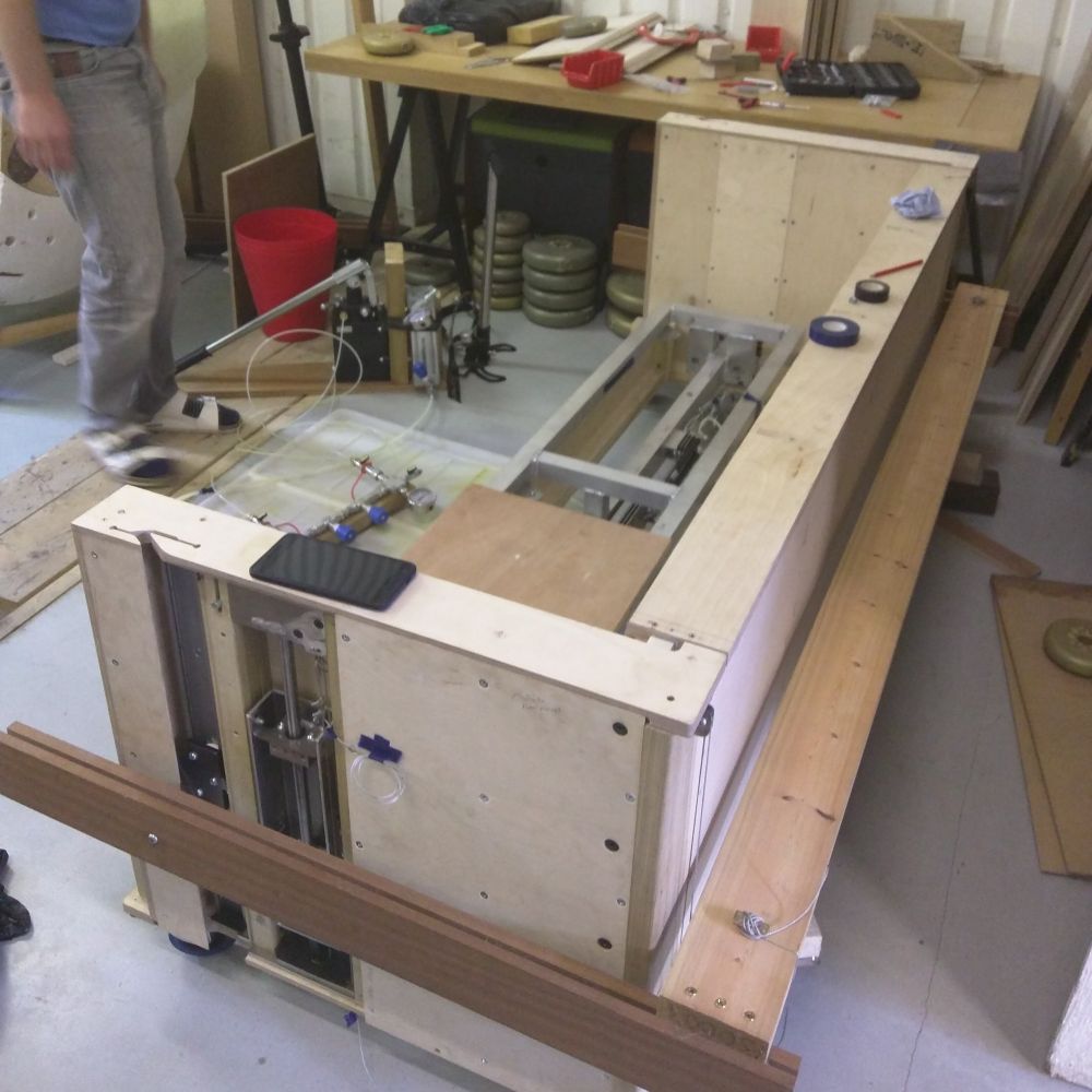 A test rig prototype in progress, constructed from plywood, metal components, and hydraulic elements, designed for exploring multiple ideas in a hands-on, iterative process. The surrounding workspace is filled with tools, materials, and components, reflecting an active design exploration. This prototype serves as a tangible way to engage with design narratives—testing not just functionality, but also interactions, material behaviours, and potential user experiences. The iterative nature of this setup aligns with the ethos of the Materials Intelligence Lab, where experimentation informs deeper material and design understanding.