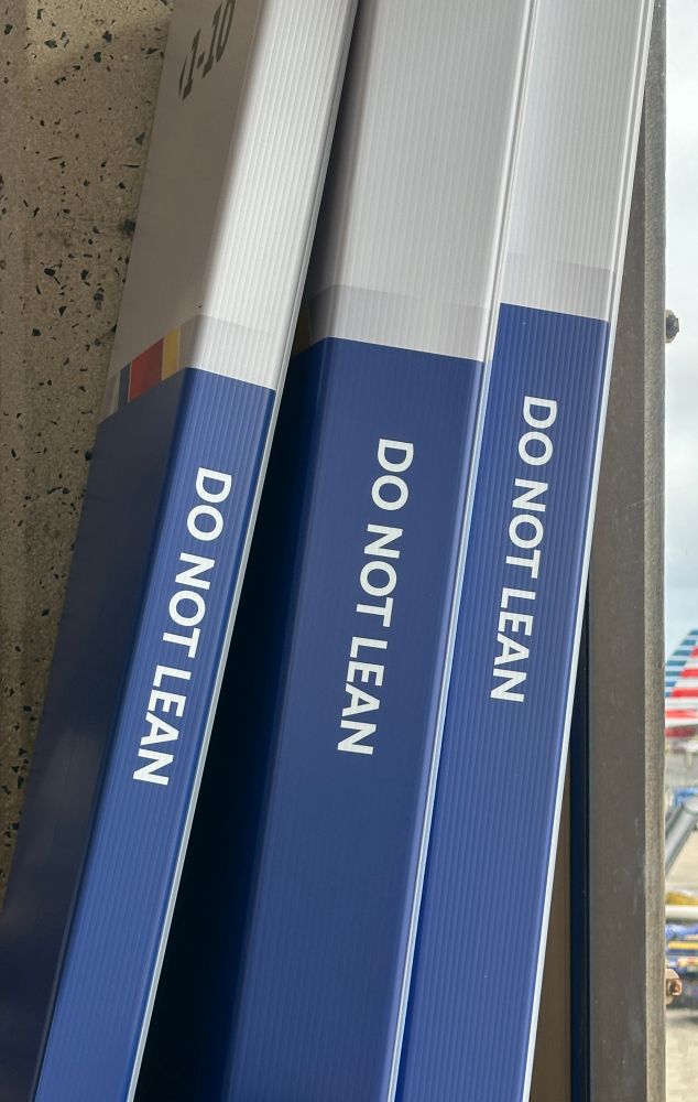 Photo of three tall slim airline packing boxes - maybe for skis? - in front of a square concrete pillar, leaning to the right against a terminal window. Through the skin sliver of glass, part of an airplane tail is visible. 