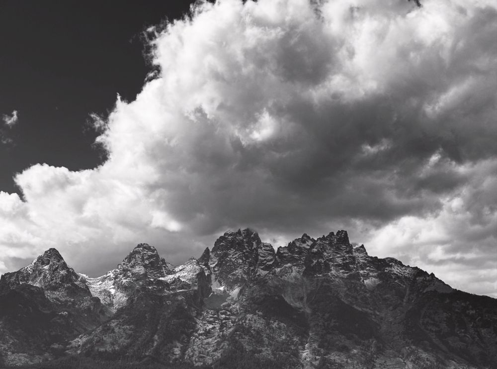 Black and white photo of four slightly hazy main peaks in the Grand Teton Mountain Range, craggy and gray with dustings of snow at the top. The sky is filled with large dramatic puffy clouds, except for a clear dark gray area in the upper left.