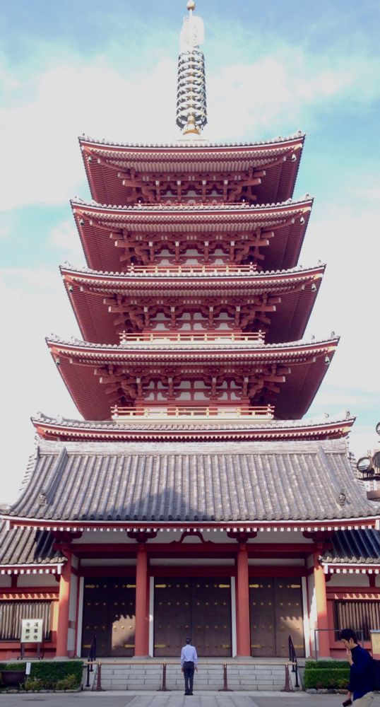 Washed-out photo looking straight at the central section of a 5-story Buddhist temple with a broad tile roof above the entrance and showing the red undersides of the upper levels, with ornate wooden framing, all topped by a golden spire. At the base, a man stands stiffly in the middle distance looking up at the temple in front of him; a young man is just entering the frame at the bottom right, striding purpusefully as he holds his phone in one hand and puts his index finger to his lips in thought with the other.