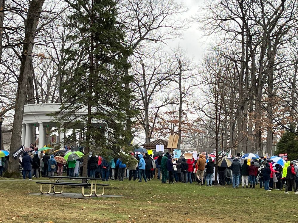 View from a distance of hundreds of people gathered around a park gazebo in the rain.