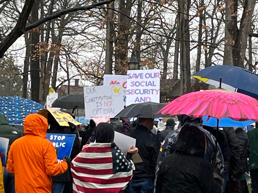 Closeup of protesters and raincoats with umbrellas. Signs say “Save Our Social Security” and “Our Constitution is not Optional.”