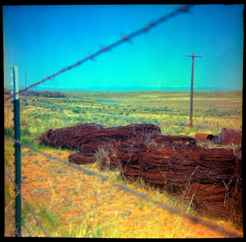 Rolls of barbed wire are stacked behind a barbed wire fence. The land around it is flat and dry and spotted with green shrubs. The sky is cloudless. 