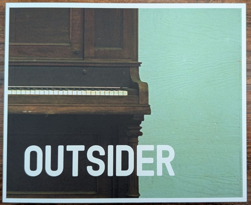 The cover of the book outsider. The photo is a close up of the right half of a piano in front of a painted plywood wall.