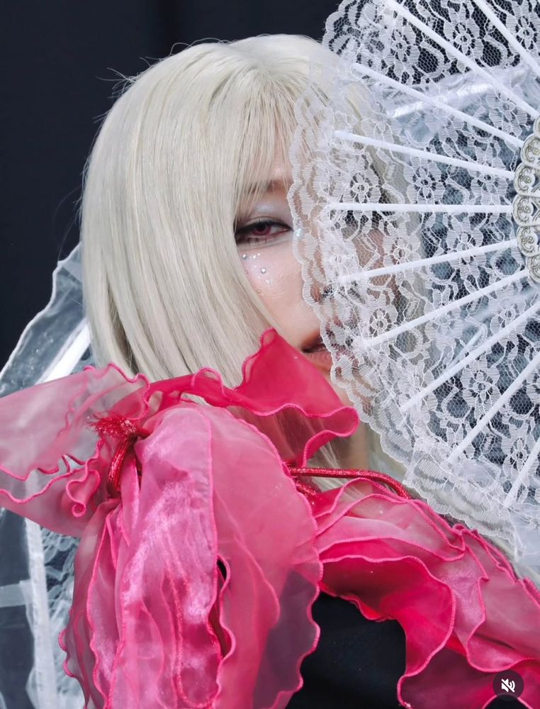 Rui of xlov with a white fan covering half his face, long white hair, and a black top with red and pink ruffles. 