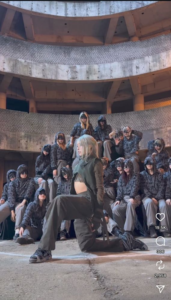 Rui kneeling on one leg, arms straight down his sides. Behind him are background dancers. All are in grey/black outfits, Rui's is a crop top. The location is like a concrete collosusm.