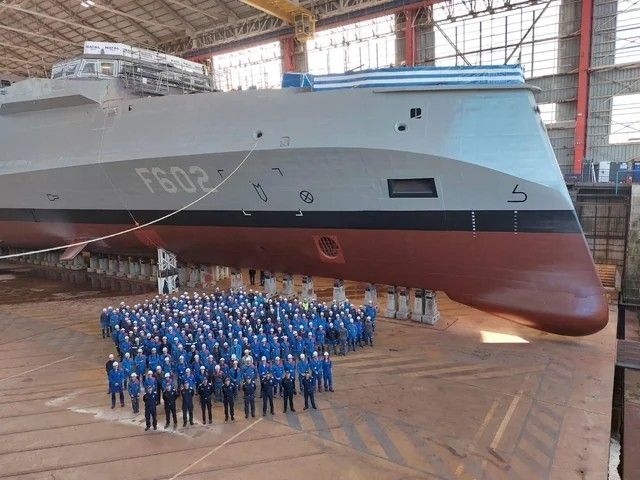 Bunch of people in hard hats with a new Greek frigate behind them, still under construction. The frigate appears to have an extraordinary chin...