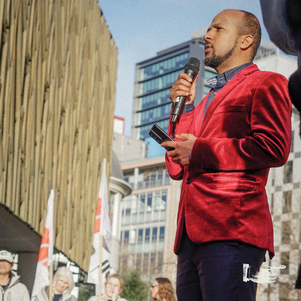 A photo of nureddin mohamed abdurahman speaking in front of the supreme court, looking incredible in a red suede coat. people in the background are holding workers first union flags