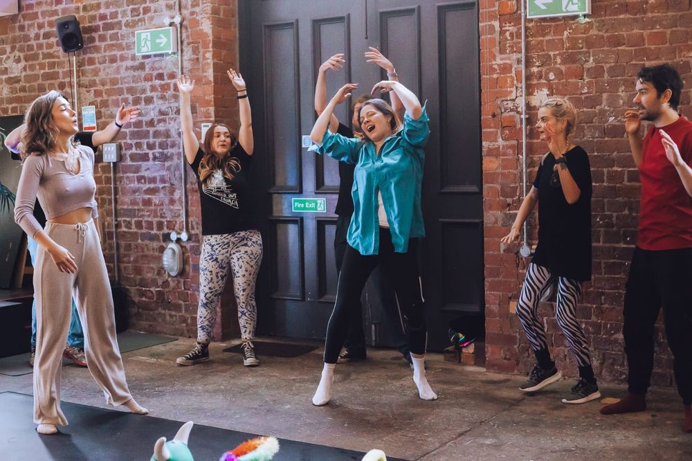 A group of people in leggings, trackies and tshirts, dance, wave arms above heads, and look happy to be starting their day.
